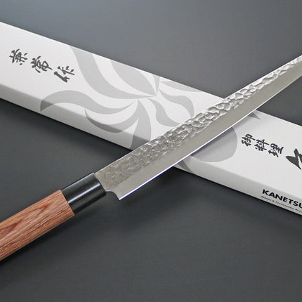 KC-959 Sujihiki Carbon 240mm Kanetsune knife with hammered blade and wooden handle beside its packaging box.