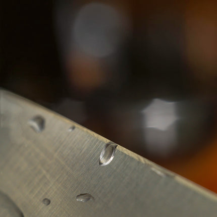 Close-up of a wet, brushed metal knife blade edge, highlighting water droplets and the sharp bevel. Razor Sharp AZ.